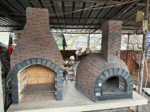 Bir atölye ortamında duran, solda ızgaralı barbekü, sağda kubbeli taş fırın içeren, el yapımı kırmızı ve gri tuğlalarla kaplanmış büyük bir dış mekan pişirme ünitesi.