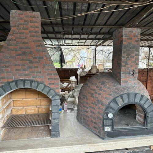 Bir atölye ortamında duran, solda ızgaralı barbekü, sağda kubbeli taş fırın içeren, el yapımı kırmızı ve gri tuğlalarla kaplanmış büyük bir dış mekan pişirme ünitesi.