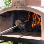 Tekerlekli metal stand üzerinde, topraklı kubbe ve bacaya sahip, odun ateşiyle çalışan modern portatif bahçe taş fırını.