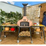 Siyah stand üzerinde, pembe/toprak rengi kubbeli, boru bacalı Tashoven Pro 100 model odunlu taş fırın. Altında odunluk ve yanlarında tekerlekli hazırlık masaları mevcuttur.